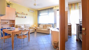 A bright living room with a sofa, a dining table with chairs, a kitchen area with a counter and shelves, and a door leading to another room.