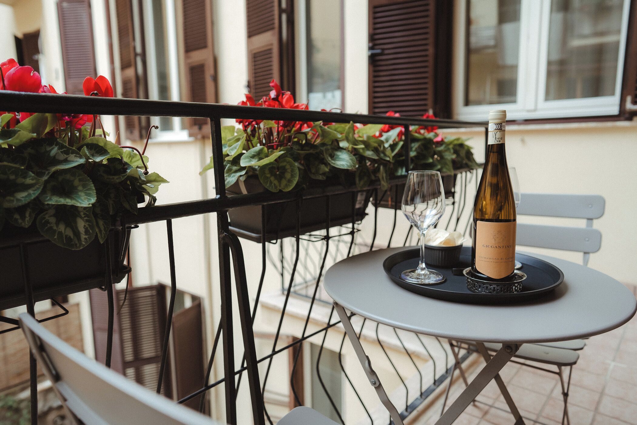 A table with a bottle of wine, two glasses, and a tray on a balcony with potted plants and a railing.