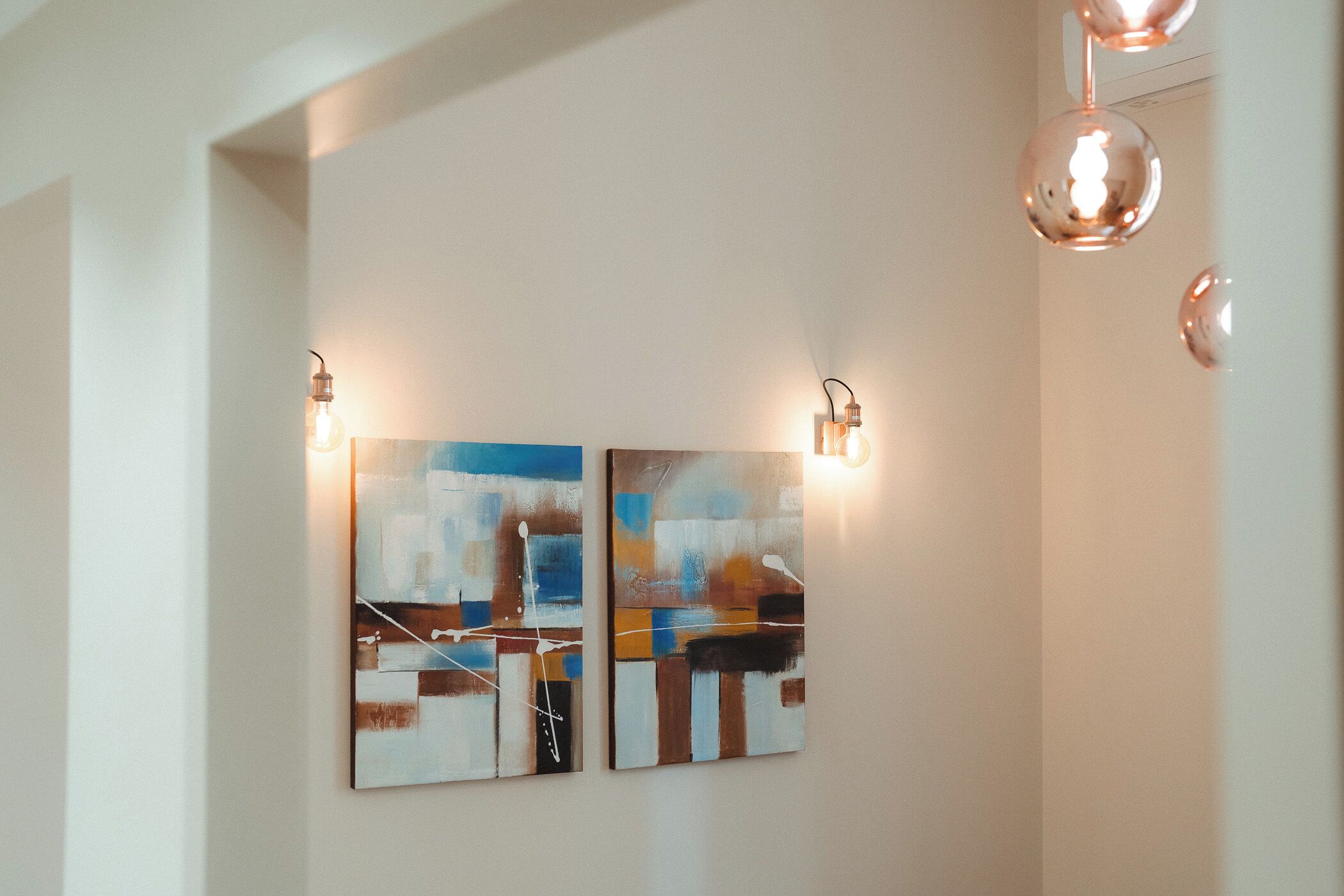 Twee abstracte schilderijen aan een muur, verlicht door wandlampen.