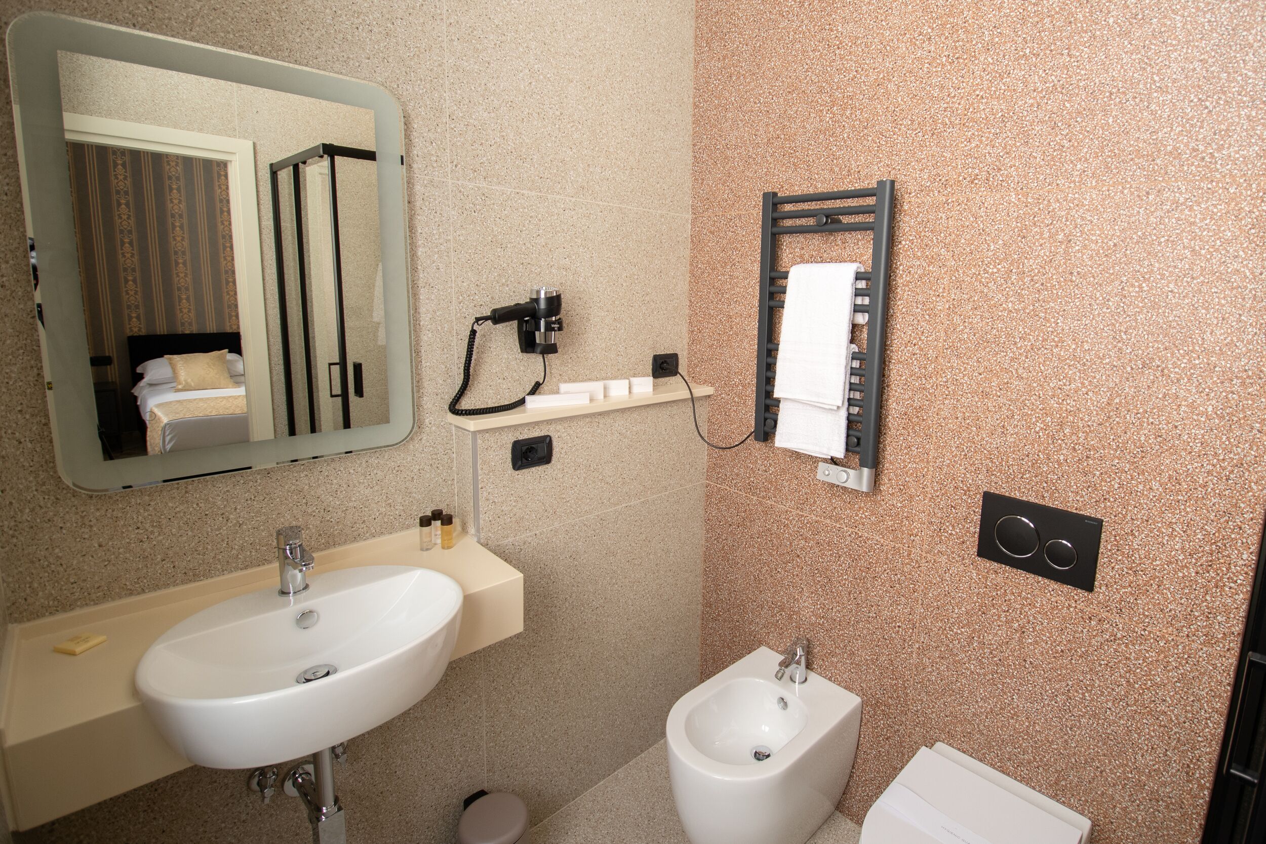 A bathroom with a sink, bidet, mirror, towel rack, and a towel dispenser.