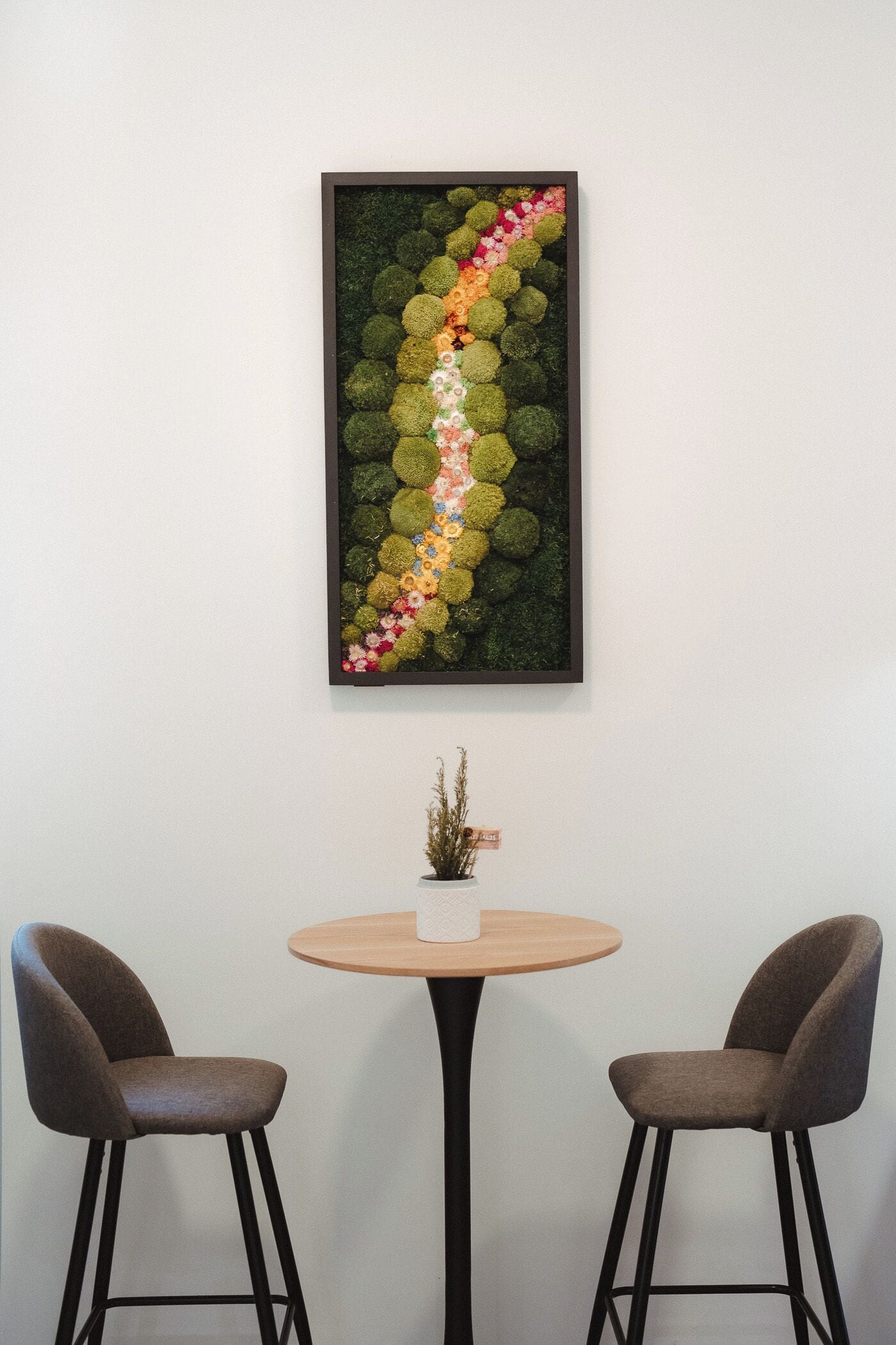 A framed artwork of a floral arrangement on a wall, a round wooden table with a small potted plant, and two chairs.