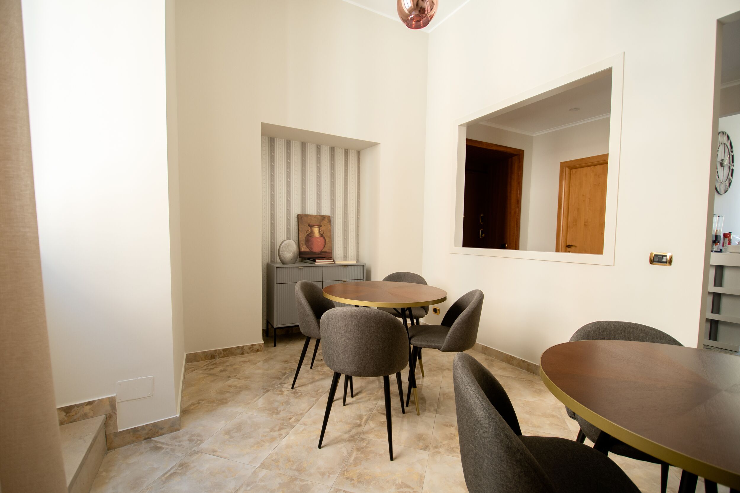Un comedor con una mesa redonda y seis sillas, un mueble pequeño con un jarrón, y un reloj en la pared.