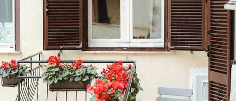 A balcony with a metal railing, a small table, and two chairs. There are potted plants with red flowers on the railing.