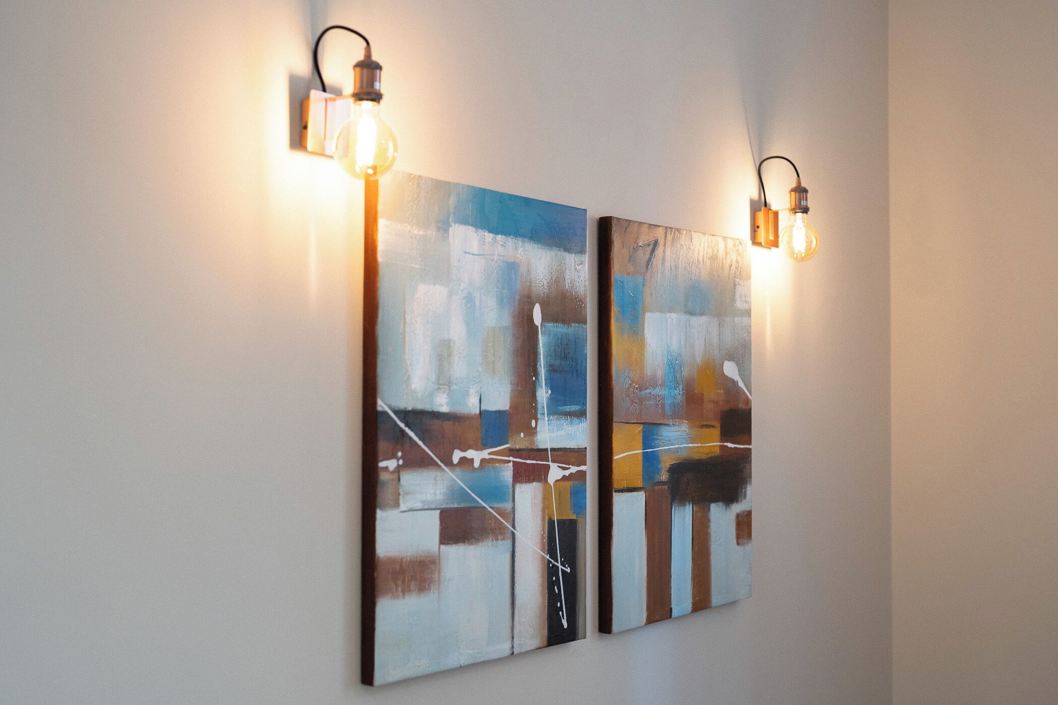 Deux tableaux abstraits sont accrochés à un mur et éclairés par des luminaires fixés au mur.