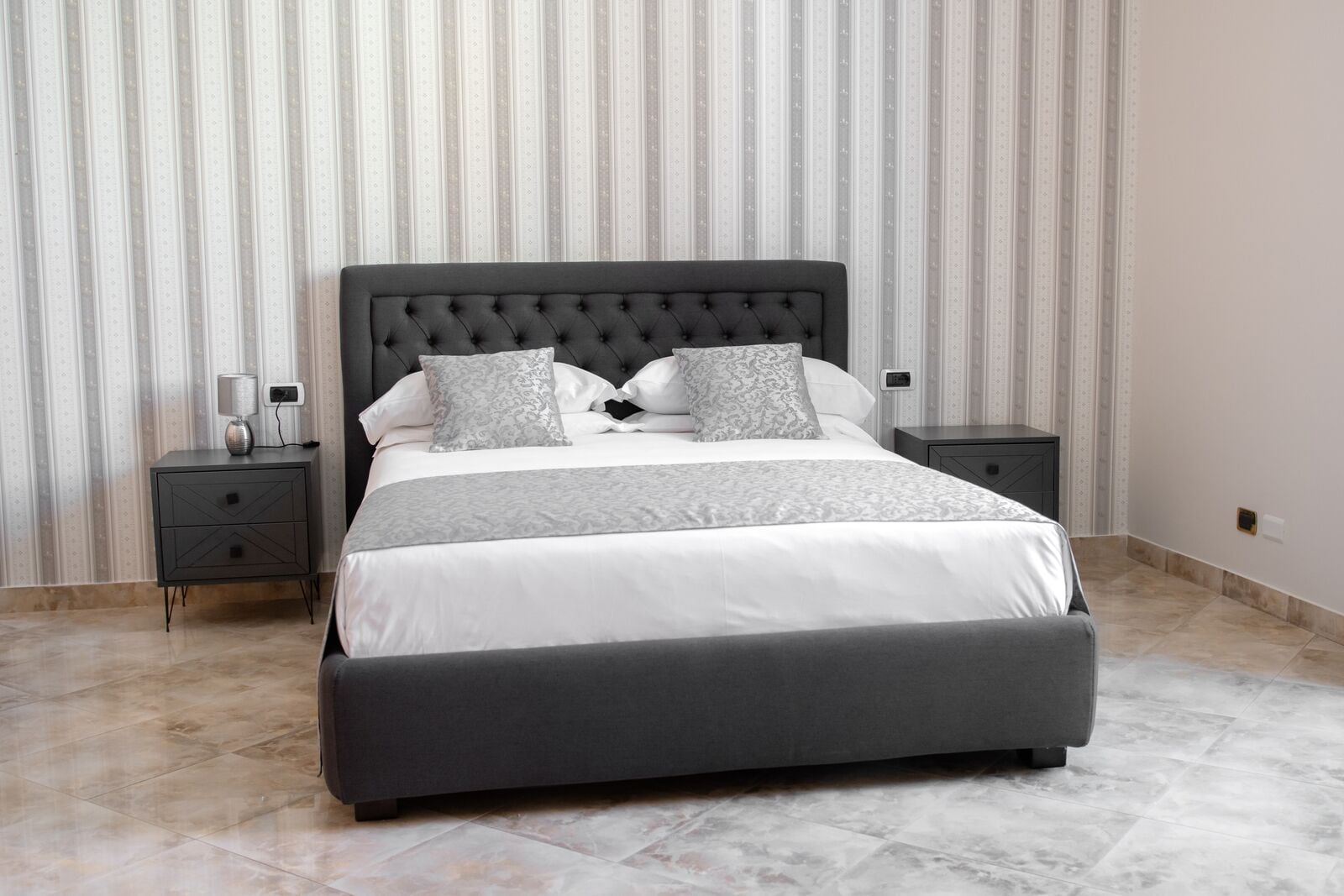 A modern bedroom with a large bed, two bedside tables, and a striped wallpaper background.