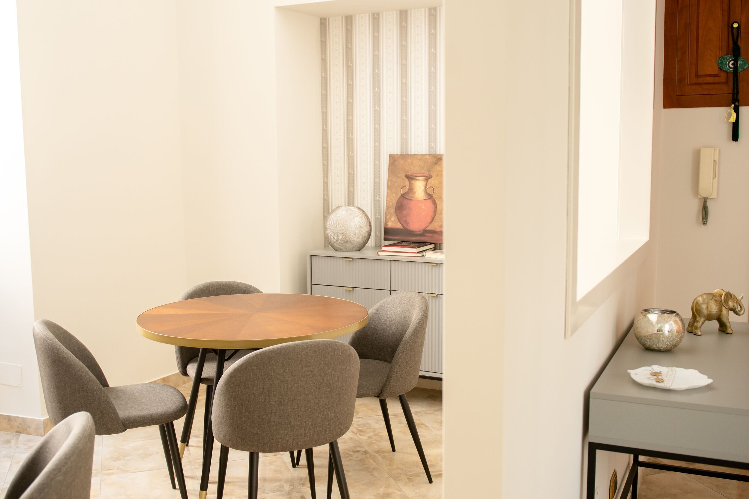 Un comedor con una mesa redonda y cuatro sillas, un aparador con objetos decorativos y un teléfono en la pared.