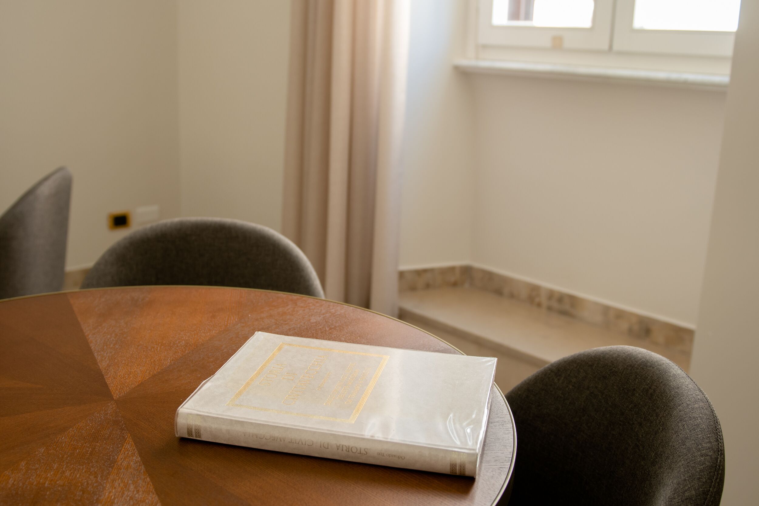 A round wooden table with a book on it, a gray chair, and a window with curtains in the background.