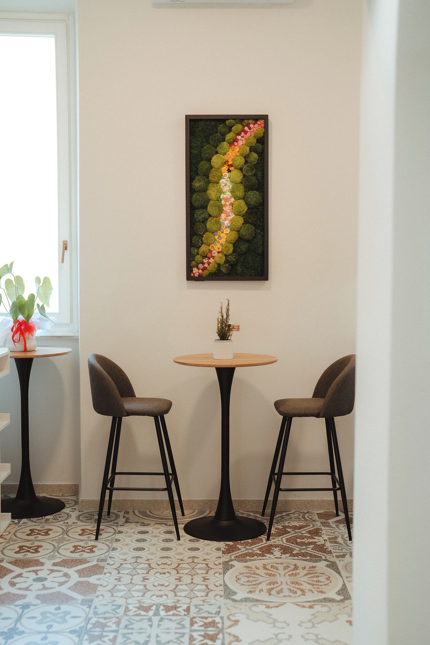A corner room with two chairs, a small table, a potted plant, and a framed artwork on the wall.