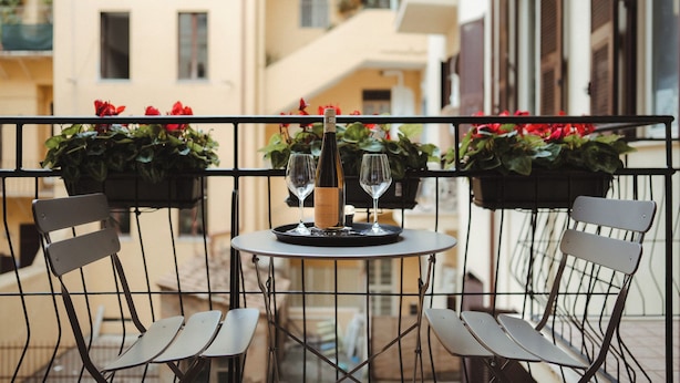 Un balcón con una mesa lista para dos personas, que incluye una botella de vino y dos copas. Hay plantas en macetas con flores rojas a cada lado de la mesa.