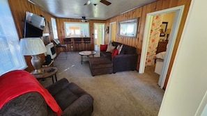 Una habitación acogedora con sofá, reposapiés y una pequeña mesa con lámpara. Hay un televisor, una ventana con persianas y una puerta con cortina.