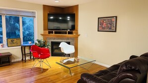 A living room with a fireplace, a television, and a large window with a view of trees. - Cozy house-3Bedrm-Aircon-2 Free parking-inner city (Calgary)