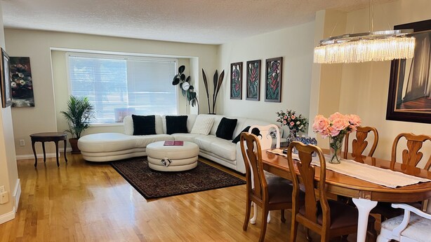 A living room with a white sofa, a dining table with chairs, a wooden floor, and a large window with blinds. - Cozy house-3Bedrm-Aircon-2 Free parking-inner city (Calgary)
