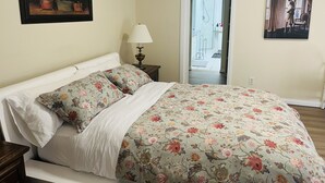 A bedroom with a bed, floral bedding, a nightstand with a lamp, a door leading to a bathroom, and a painting on the wall. - Cozy house-3Bedrm-Aircon-2 Free parking-inner city (Calgary)