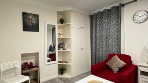 A modern living room with a red sofa, white armchair, and a small white table with a woven placemat. A white cabinet with shelves is against the wall, and a clock is visible on the wall. - Acton Vale hotel  (London)