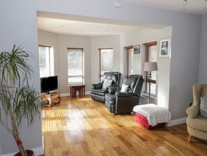 A spacious living room with a wooden floor, a leather sofa, a red ottoman, and a television on a wooden stand.