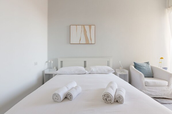 A hotel room with a large bed, white bedding, a bedside table with a lamp, a chair, and a painting on the wall.