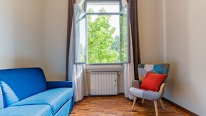 A room with a blue sofa, a chair with a patterned cushion, a window with curtains, and a radiator. - GuestHost - The apartment, a welcoming residence of approximately 70 square meters, is ideal for hosting up to 4 people. Located on the first floor of a building with elevator in the Washington district, one of the most elegant residential areas of Milan (Milano)