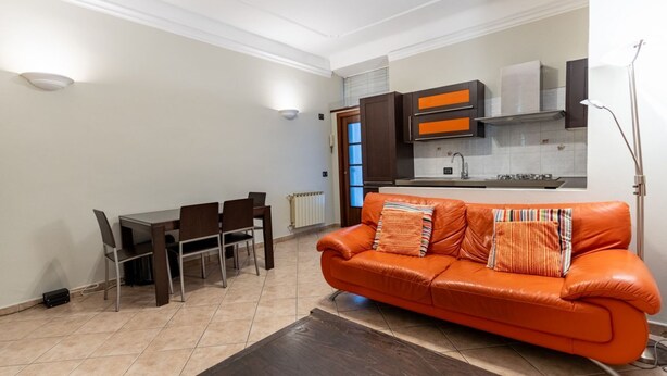A modern kitchen with dark cabinets and a window, a dining area with a table and chairs, and an orange leather sofa with pillows. - GuestHost - The apartment, a welcoming residence of approximately 70 square meters, is ideal for hosting up to 4 people. Located on the first floor of a building with elevator in the Washington district, one of the most elegant residential areas of Milan (Milano)