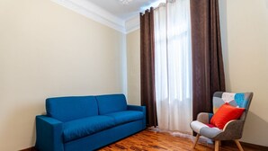 A room with a blue sofa, a grey armchair with a red pillow, wooden flooring, and a window with brown curtains. - GuestHost - The apartment, a welcoming residence of approximately 70 square meters, is ideal for hosting up to 4 people. Located on the first floor of a building with elevator in the Washington district, one of the most elegant residential areas of Milan (Milano)