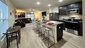 Et moderne køkken med en central ø, rustfrie hvidevarer og et spiseområde med sofa og stole.