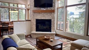 A spacious living room with a stone fireplace, a flat-screen TV mounted above it, and large windows offering a view of the outdoors. - Hot Tub & Mtn Views - Modern Build, Pet-Friendly, 14 mi to Breck (Alma)