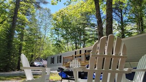 A campsite with two white Adirondack chairs, a gravel ground, and a mobile home in the background.