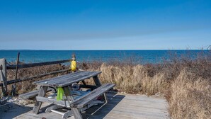 Una mesa de picnic de madera con bancos sobre una terraza de madera, con vista a una playa con hierba seca y un cielo azul despejado.