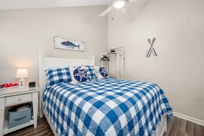 A bedroom with a checkered bedspread, a bedside table with a lamp, a clock, and a small shelf.