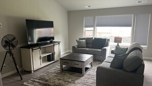 A living room with a flat-screen TV on a wooden cabinet, a grey sofa, a coffee table, and a standing fan. - Family home (Roseburg)