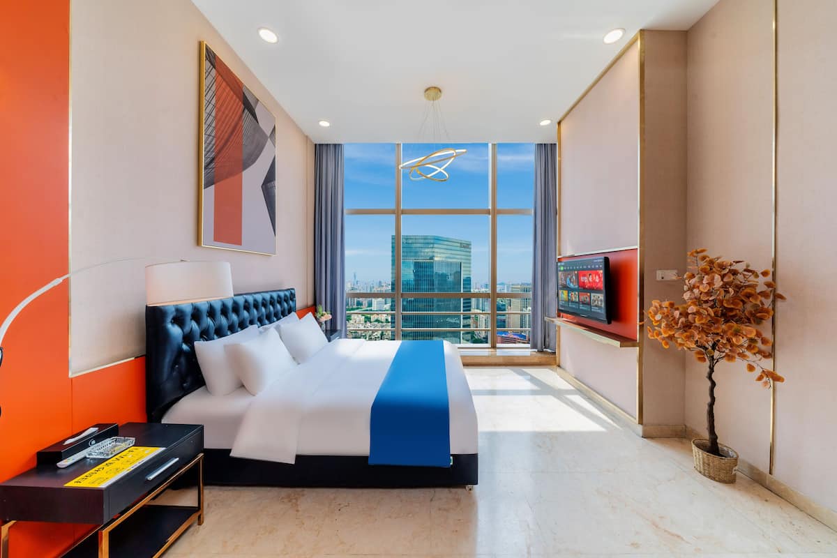 A modern hotel room with a large bed, a TV, a potted plant, and a city view through large windows.