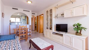 A living room with a TV, a sofa, a dining table with chairs, a kitchen in the background, and a plant on a cabinet. - Cozy apartment in Torrevieja (Torrevieja)