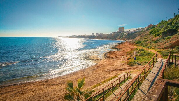 A coastal beach with a wooden boardwalk leading to the shore, a clear sky, and buildings in the distance. - Cozy apartment in Torrevieja (Torrevieja)