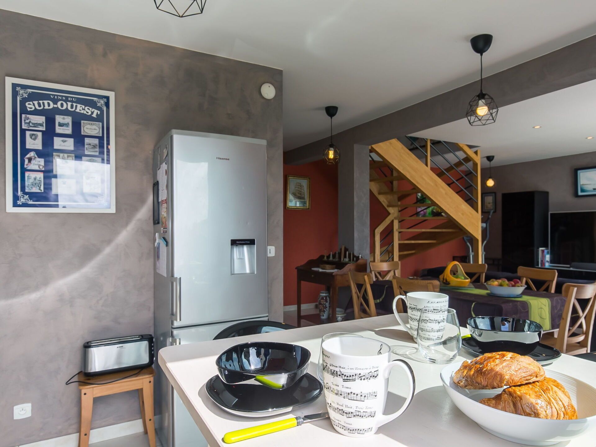 Una cocina moderna con un refrigerador, un tostador, una mesa de comedor con un bol de croissants, una taza con notas musicales y un póster en la pared.