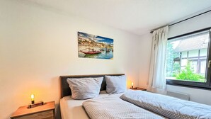 A double bed with patterned bedding, a nightstand with a lamp, a painting of a coastal scene, and a window with a view of greenery. - Cosy apartment in villa for 4 people with WIFI, TV, terrace and pets allowed (Bad Wildungen)