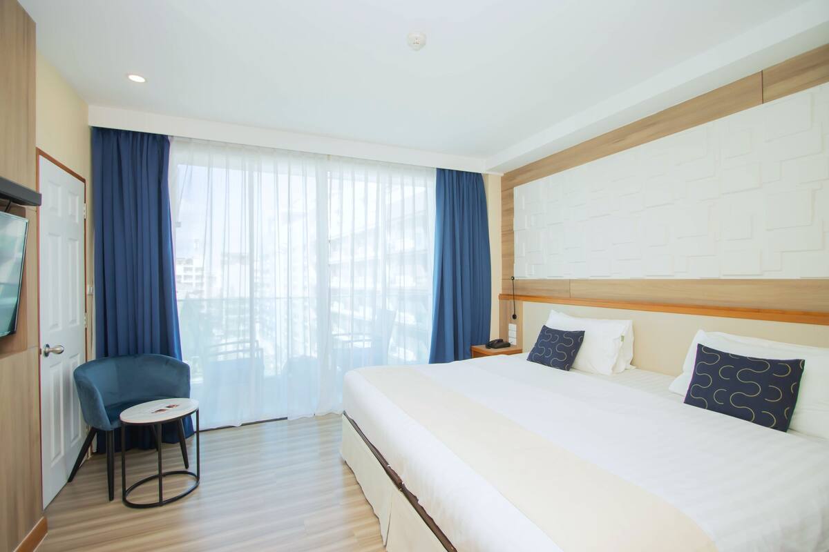 A hotel room with a large bed, a small round table, a chair, a TV, and a window with curtains.