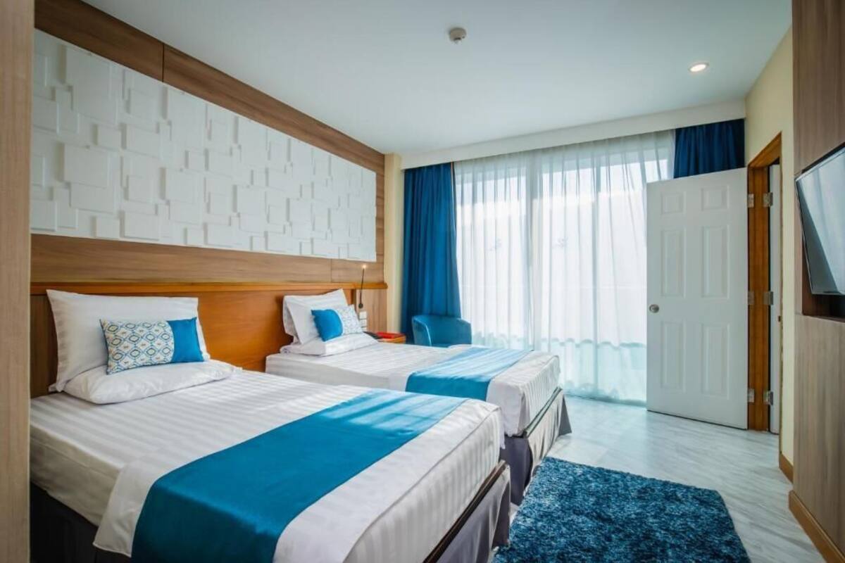 A hotel room with two beds, a wooden headboard, a large window with curtains, and a flat-screen TV.
