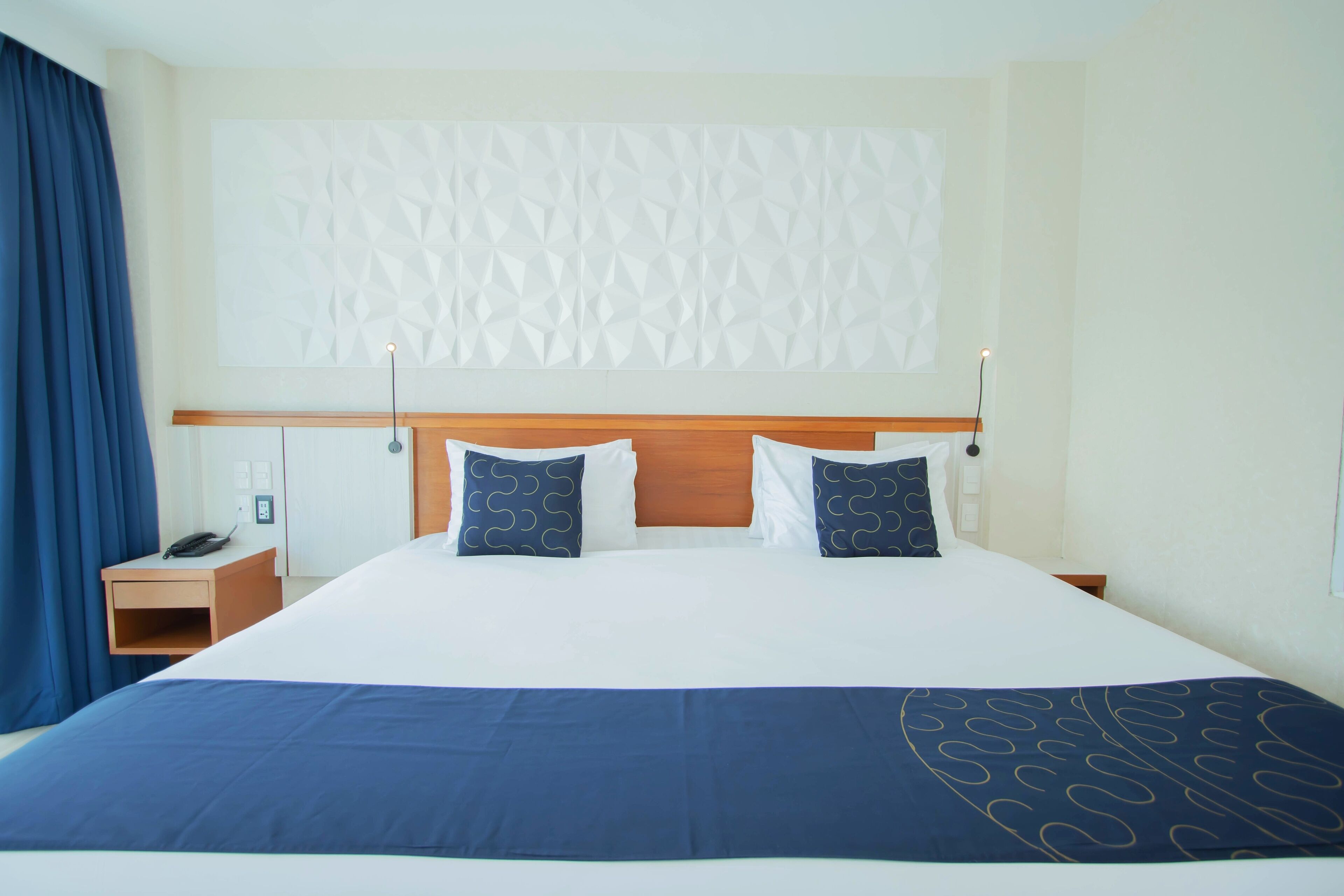 A hotel room with a large bed, a wooden headboard, two bedside tables, a nightstand, and a wall-mounted light fixture.