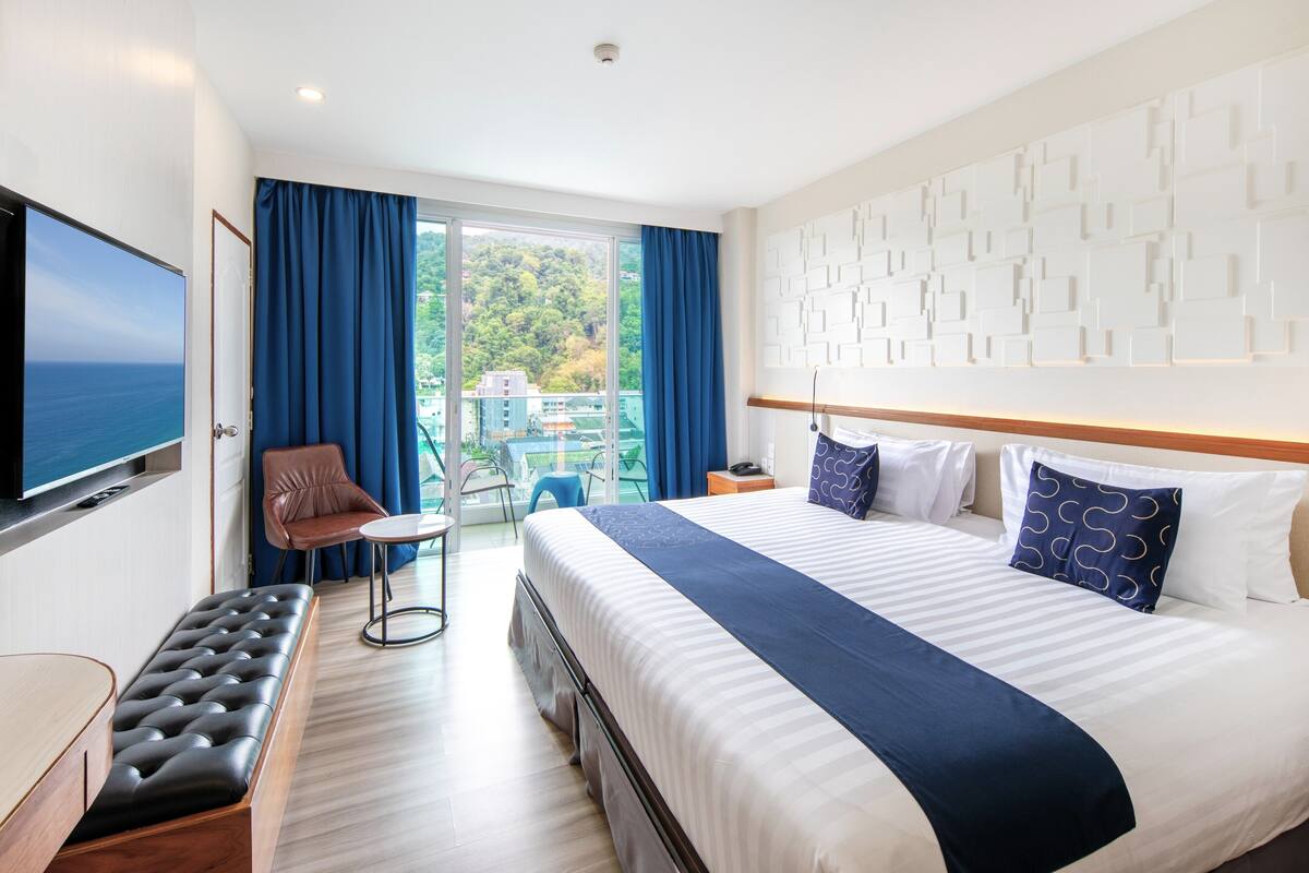 A hotel room with a large bed, a flat-screen TV, a seating area with a chair and table, and a balcony with a view of the sea.