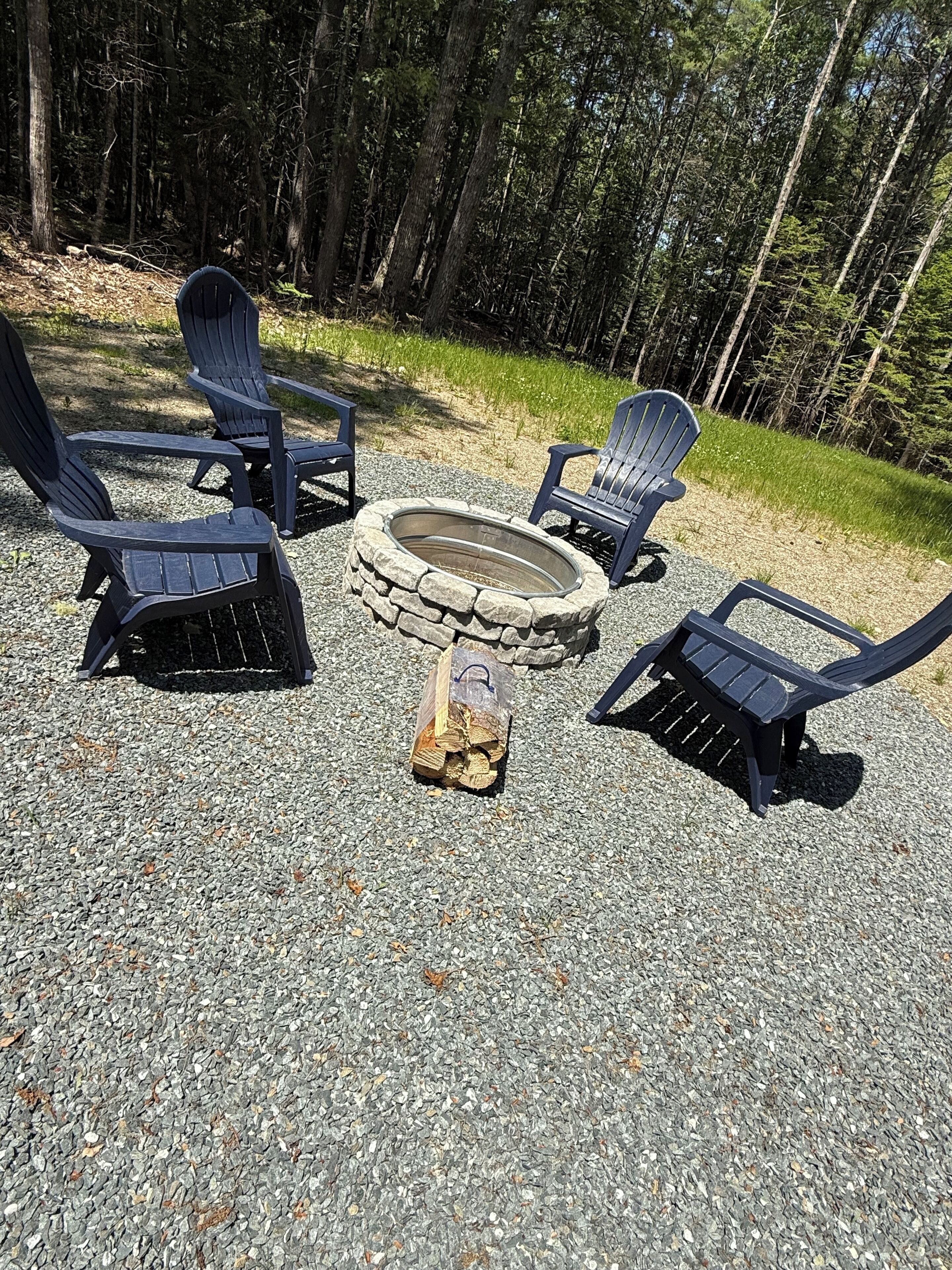 A fire pit surrounded by four Adirondack chairs on a gravel surface, with a bag of firewood in the center.