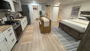 A modern kitchen with white cabinets, a stove, and a sink. There is a small dining area with a bench and a plant. A bedroom with a bed and a window is visible in the background.