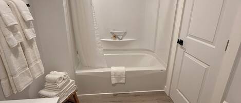 A bathroom with a bathtub, a toilet, and a sink. There is a towel rack with folded towels, a shower curtain, and a small shelf with toiletries.