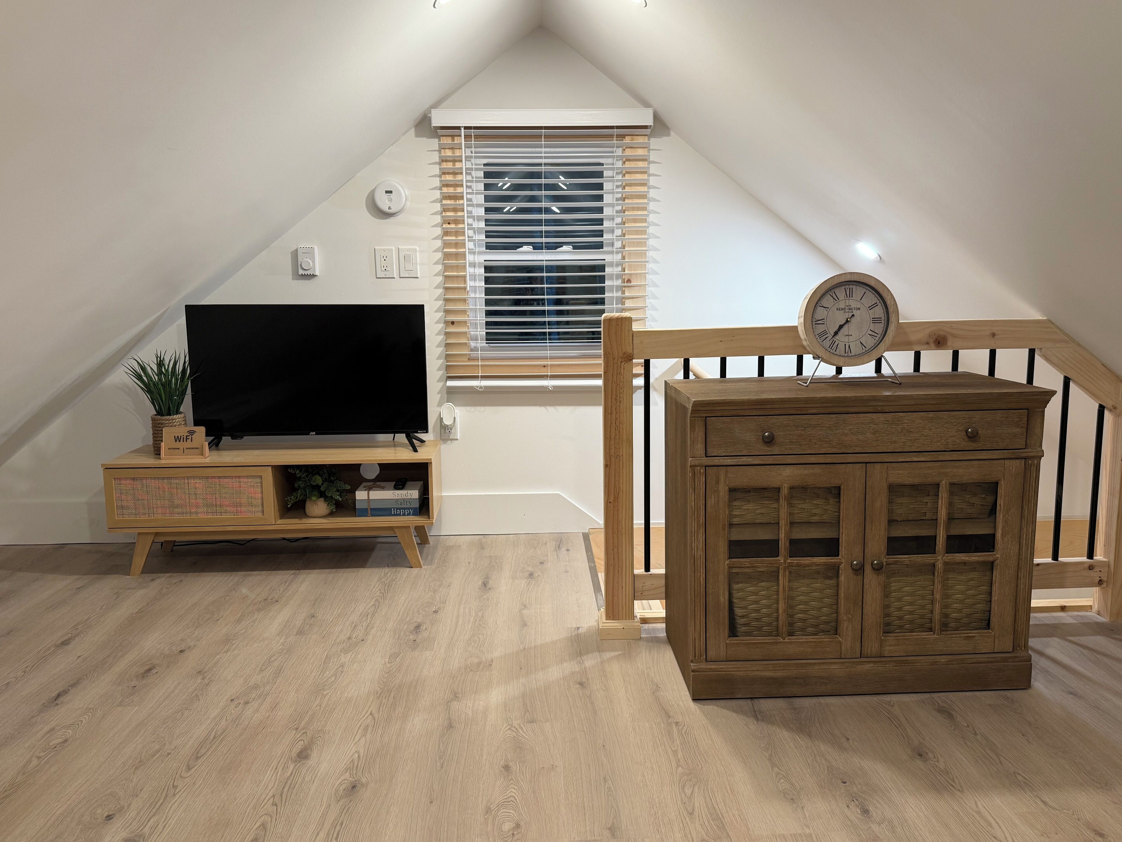 A room with a sloped ceiling, a television on a wooden stand, a clock on a cabinet, and a window with blinds.