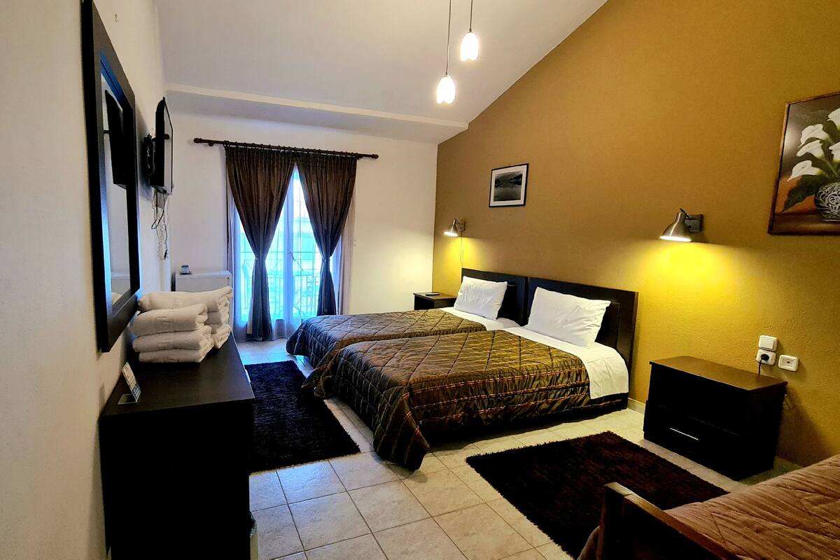 Habitación de hotel con dos camas, un escritorio, una silla, un ventanal con cortinas y un cuadro en la pared.