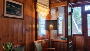 Interior de fusta amb zona de menjador, cadires, taula amb una planta, quadre amb marc i finestra que mostra una escala i arbres.