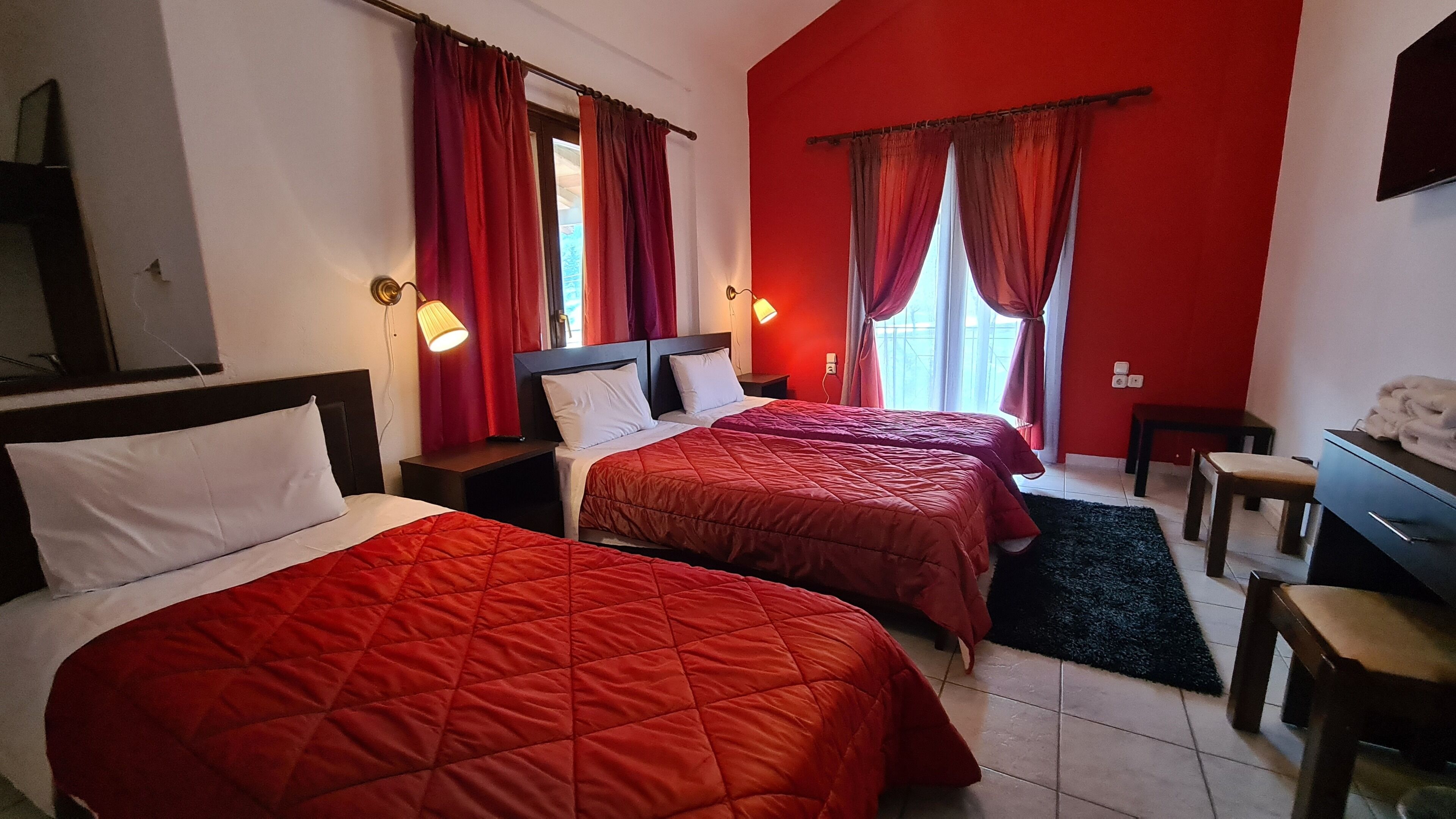 A hotel room with two beds, red curtains, a desk, and a chair.