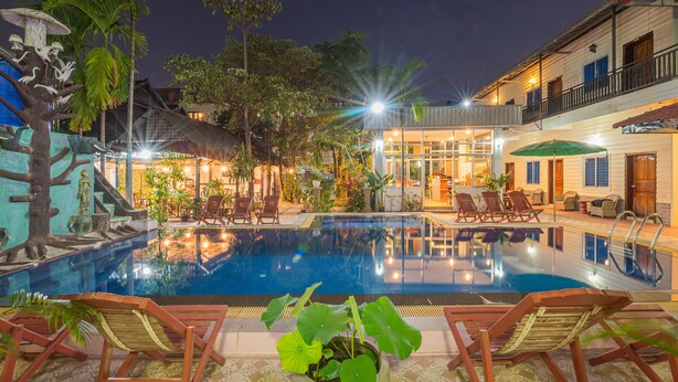 A swimming pool with lounge chairs, surrounded by tropical plants and a building with a balcony. - Lotus Bleu Guest House (Siem Reap)