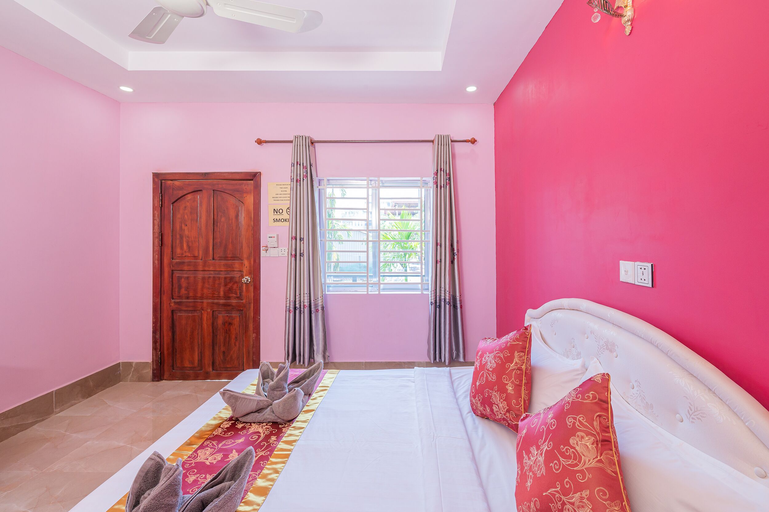 A hotel room with a bed, a window with curtains, a wooden door, and a ceiling fan.