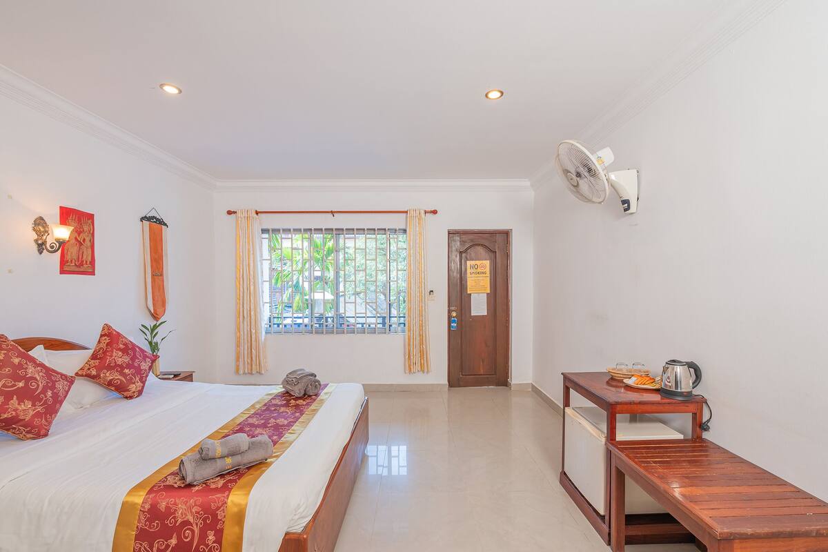 A hotel room with a bed, a small table with a kettle, and a window with curtains.