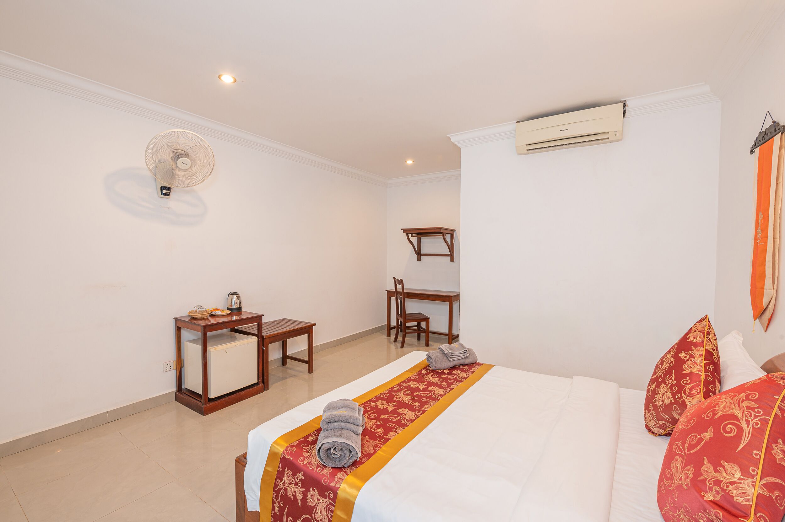 A hotel room with a bed, a desk, a chair, a fan, and an air conditioning unit.