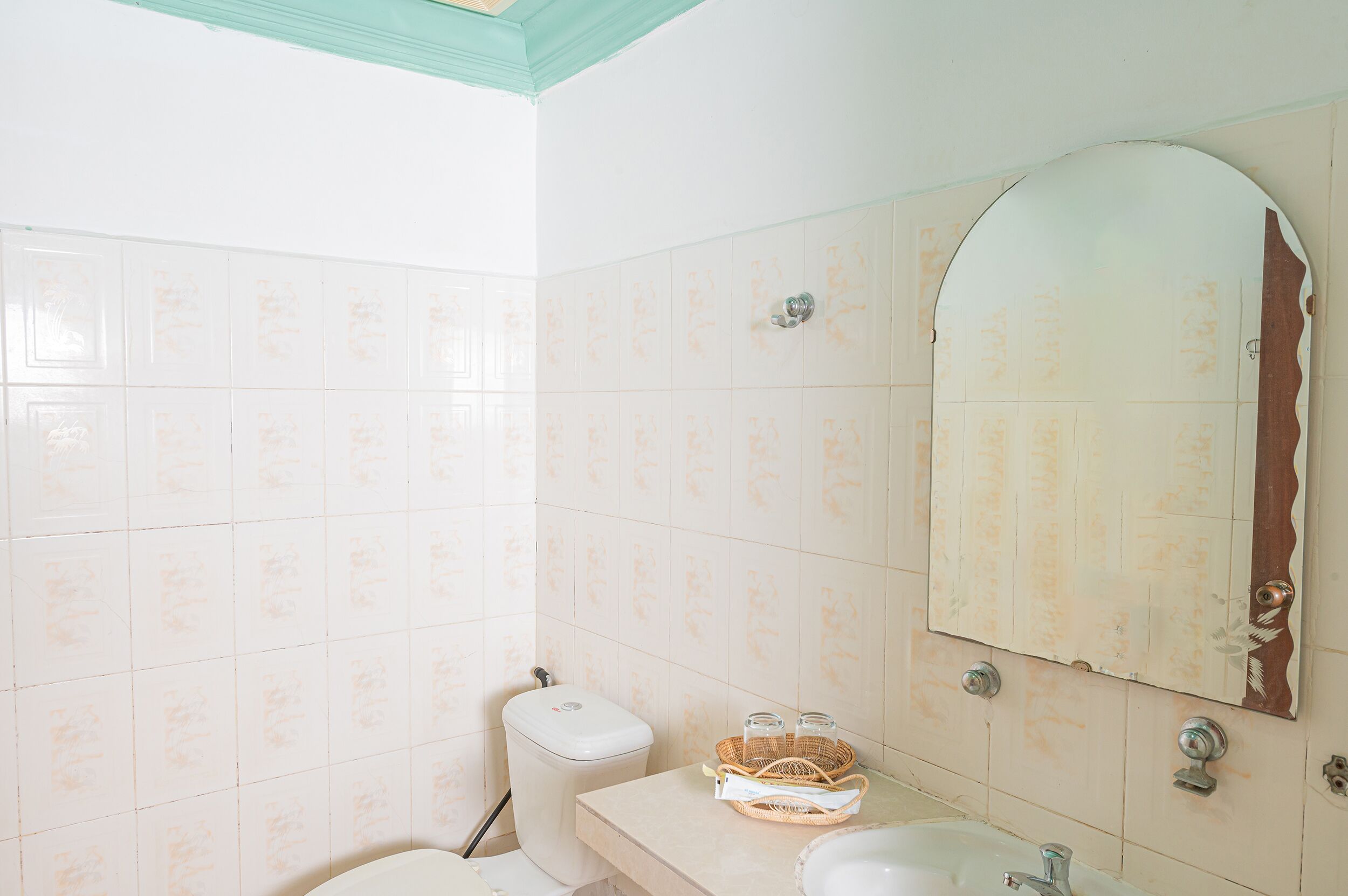 Un baño con paredes de azulejos blancos, un inodoro blanco, un lavabo con espejo y una cesta en la encimera.